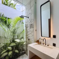 Modern open-concept bathroom with a glass-enclosed shower, white stone vanity, black-framed mirror, tropical plants, and natural stone wall.