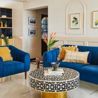 Elegant living room with blue velvet sofas, framed botanical artwork, decorative wall molding, and a patterned round coffee table.