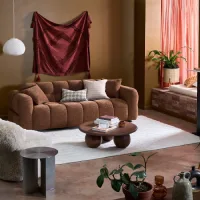 Warm living room with a brown sofa, burgundy wall hanging, white rug, wood coffee table, plants, and coral curtains