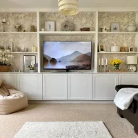 White built-in living room shelves surrounding a television, styled with framed artwork, decorative vases, flowers, and floral wallpaper above white cabinets.