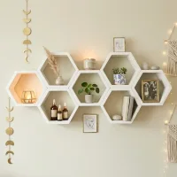 White hexagon floating shelves arranged in a honeycomb pattern on a beige living room wall, decorated with potted plants, candles, framed art, books, and warm fairy lights.