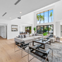 Bright open-concept living room with a large sectional sofa, black leather chairs, wood flooring, and floor-to-ceiling windows overlooking palm trees.