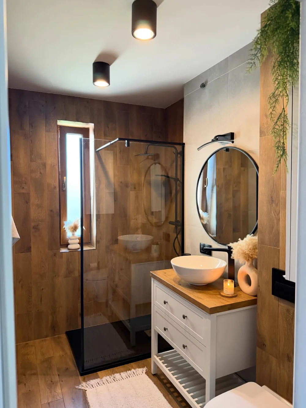 small bathroom with warm wood walls, glass shower enclosure, white vanity, and round mirror