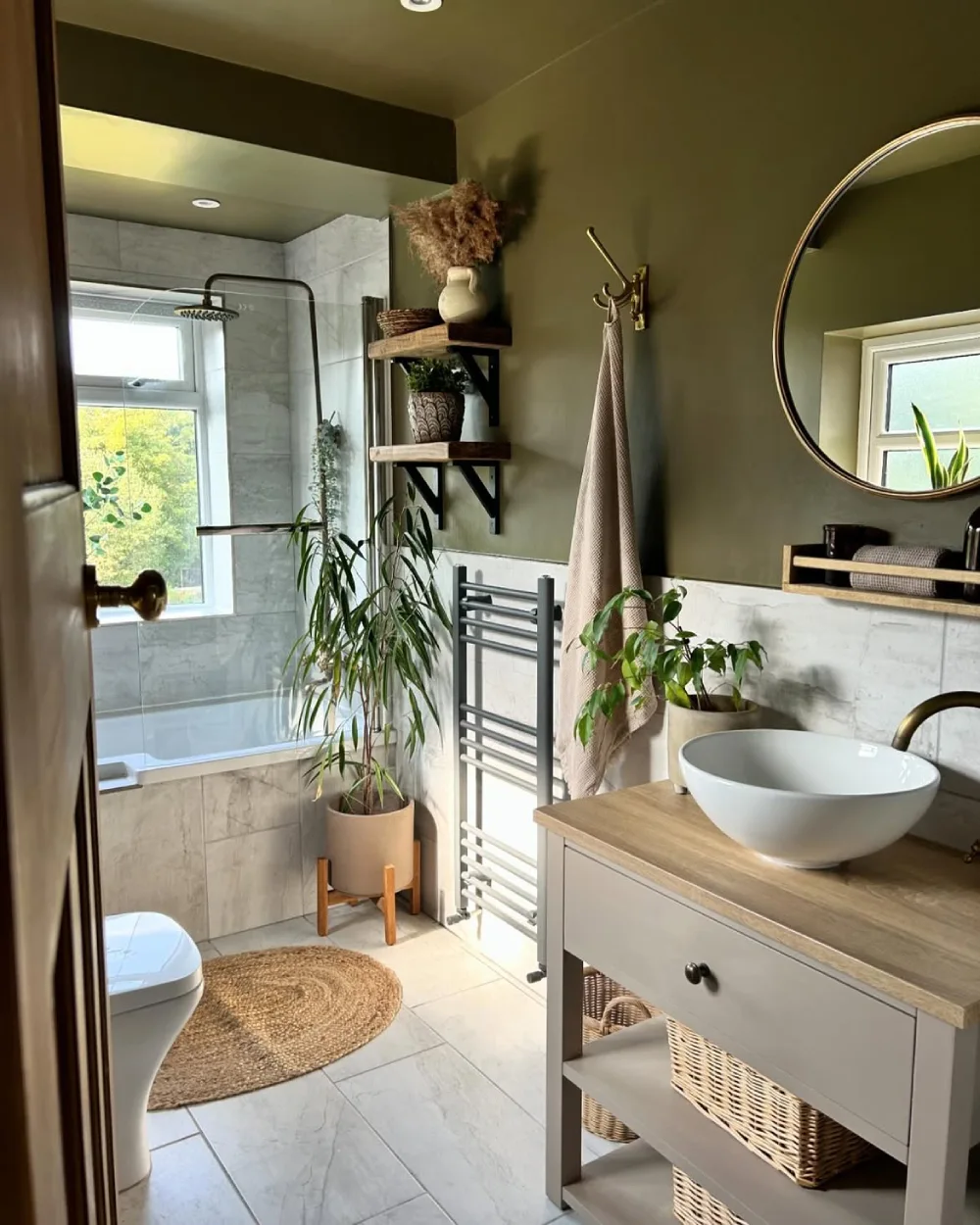 small bathroom with olive green walls, open wooden shelves, plants, and a compact vanity with woven baskets