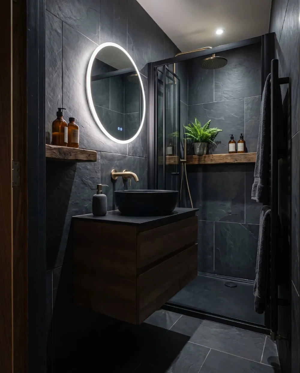 small dark bathroom with slate-look tile, floating wood vanity, black sink, and backlit round mirror