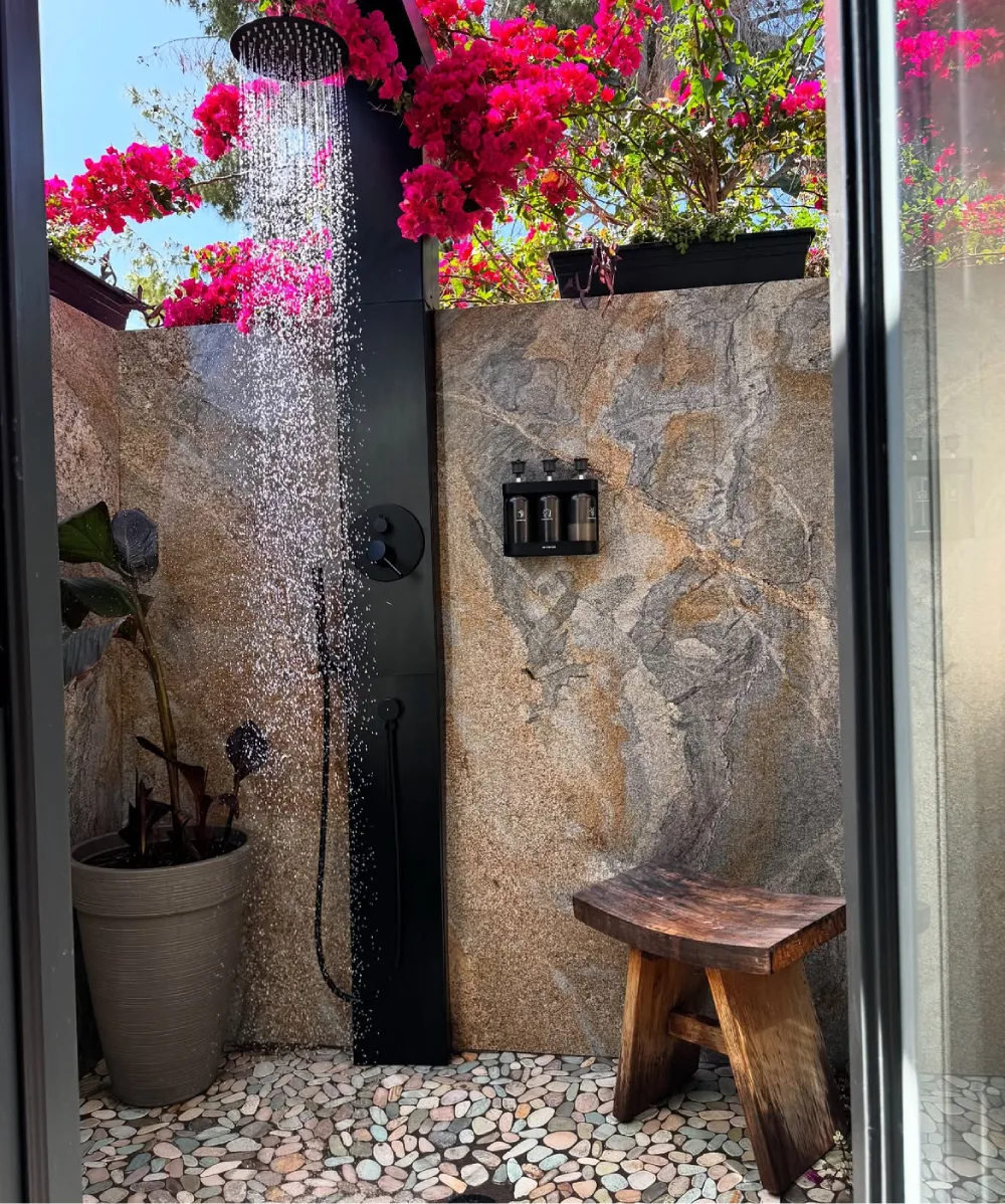 open-air outdoor shower with natural stone walls, pebble floor, tropical plants, and bougainvillea