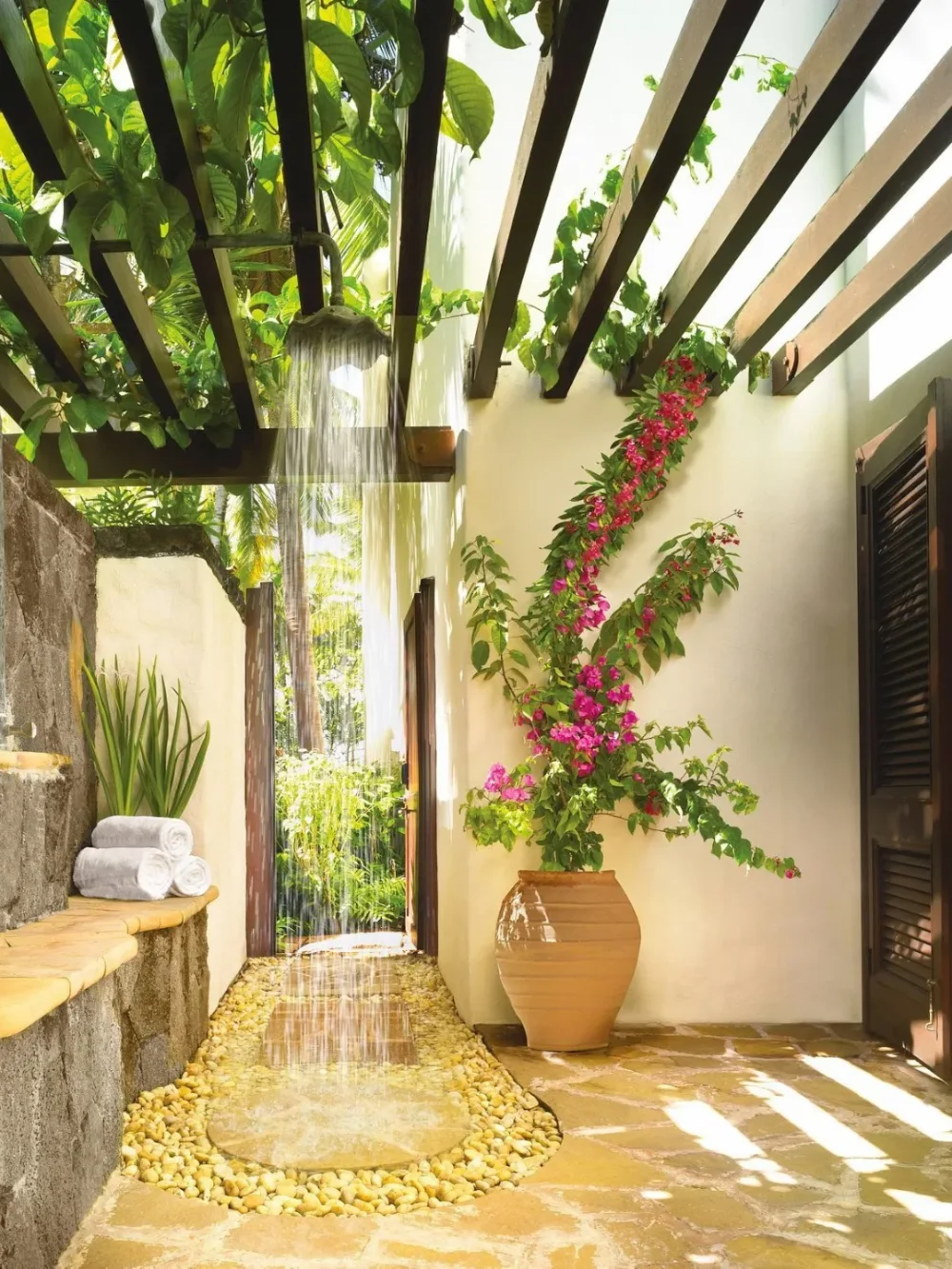 open-air bathroom with rainfall shower, wooden pergola, tropical plants, and pebble floor