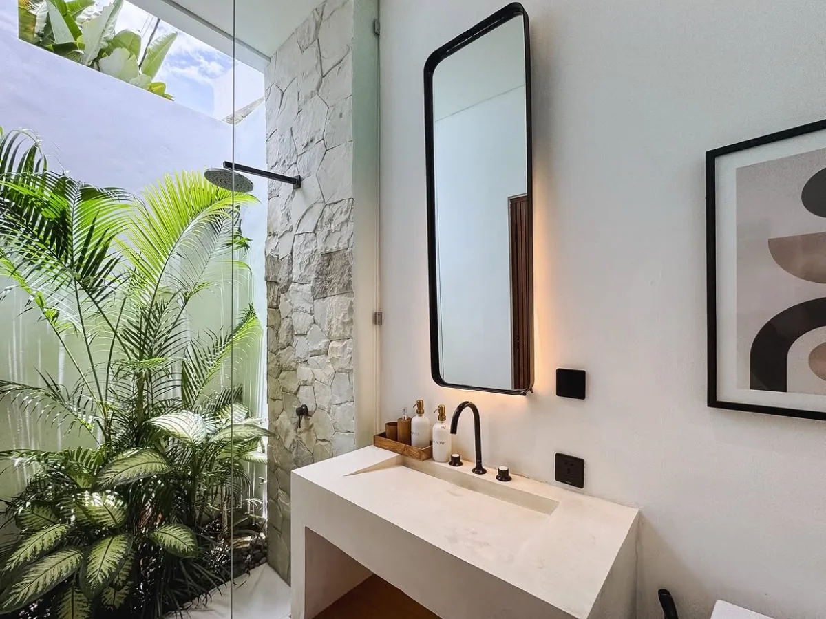 Modern open-concept bathroom with a glass-enclosed shower, white stone vanity, black-framed mirror, tropical plants, and natural stone wall.