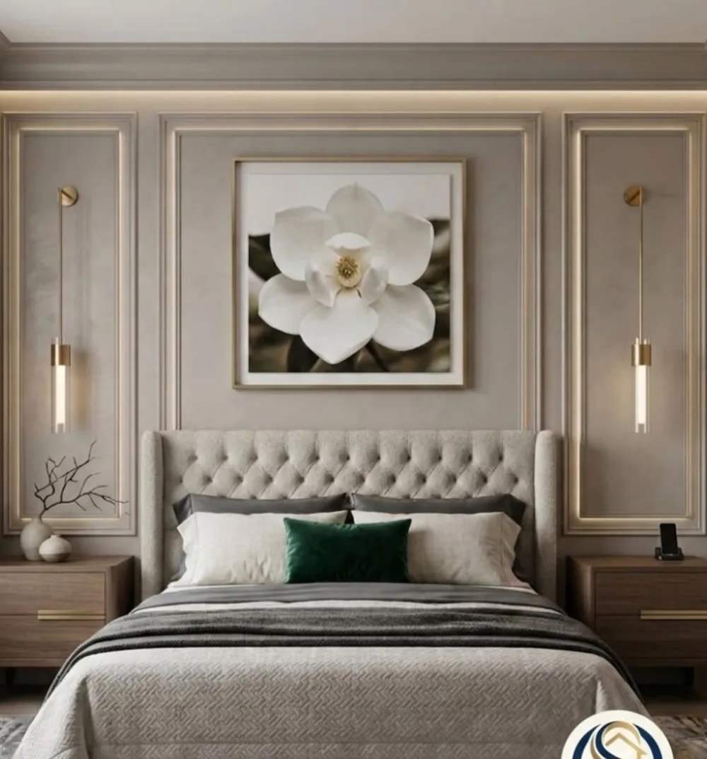 Elegant taupe bedroom accent wall with decorative molding, a tufted upholstered headboard, white floral artwork, and brass wall sconces
