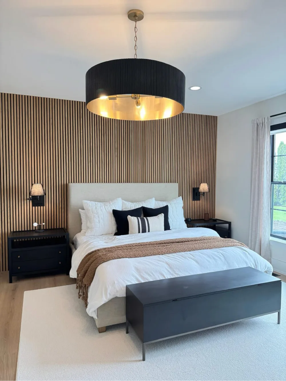 Modern bedroom with a natural wood slat accent wall behind a cream upholstered bed, black bedside furniture, and a large black drum pendant light