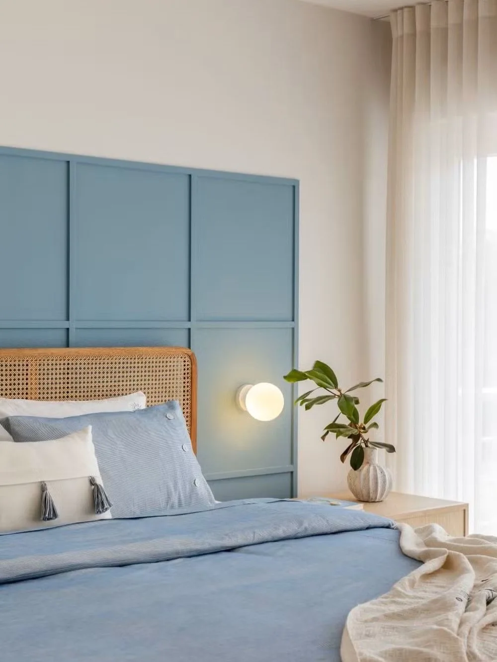 Airy bedroom with a light blue paneled accent wall, woven rattan headboard, white curtains, and soft blue bedding