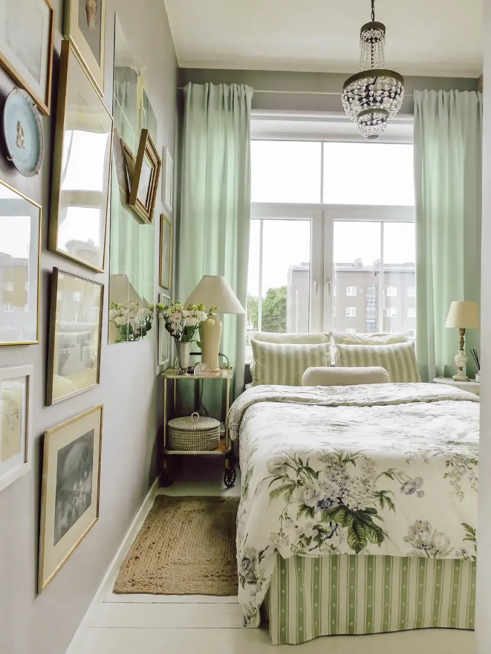 Narrow bedroom with a floral bed, slim side table, large window, and soft green curtains