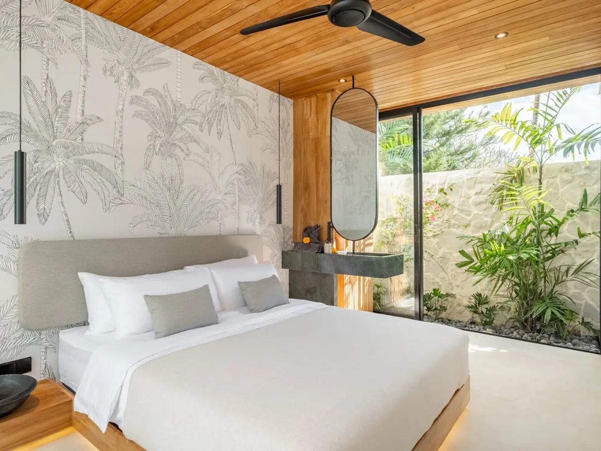 Open-concept bedroom with a tropical palm wallpaper accent wall, wood ceiling, and glass doors overlooking a private garden.