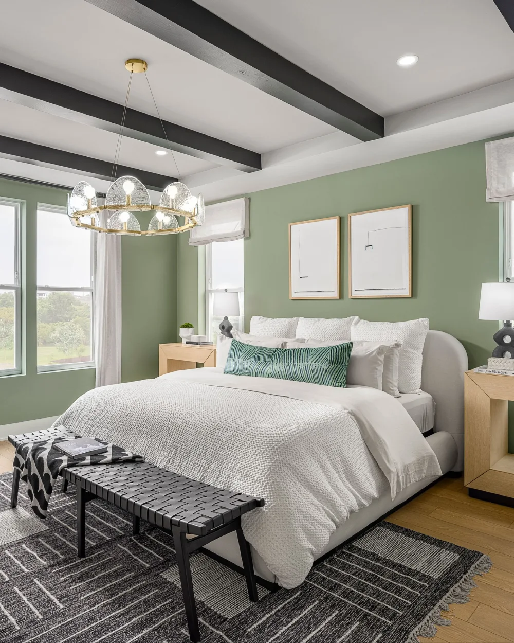 Sage green bedroom walls paired with white bedding, a gray upholstered bed, natural wood nightstands, and a black patterned rug.