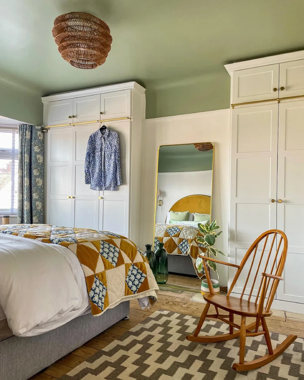 Sage green bedroom with white wardrobes, mustard and blue patchwork bedding, and a wooden rocking chair