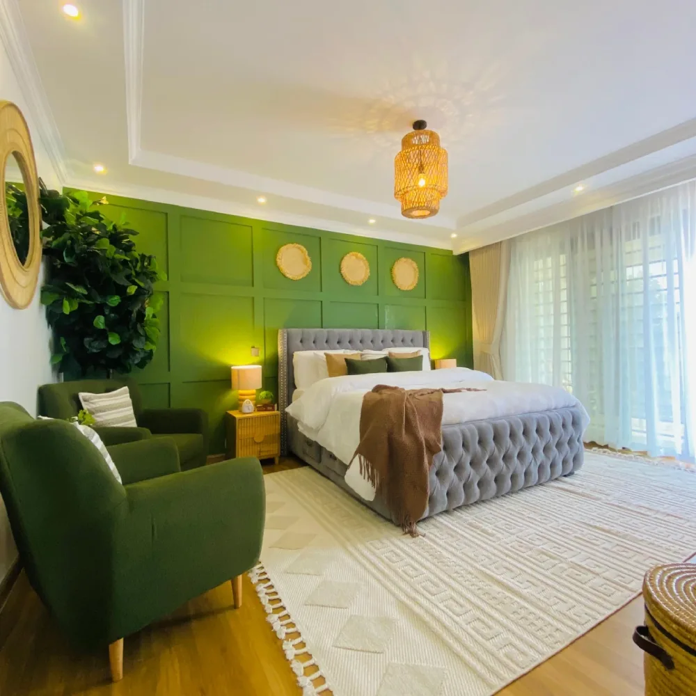 Bedroom with emerald green paneled walls, gray tufted bed, green armchairs, cream rug, and woven pendant light