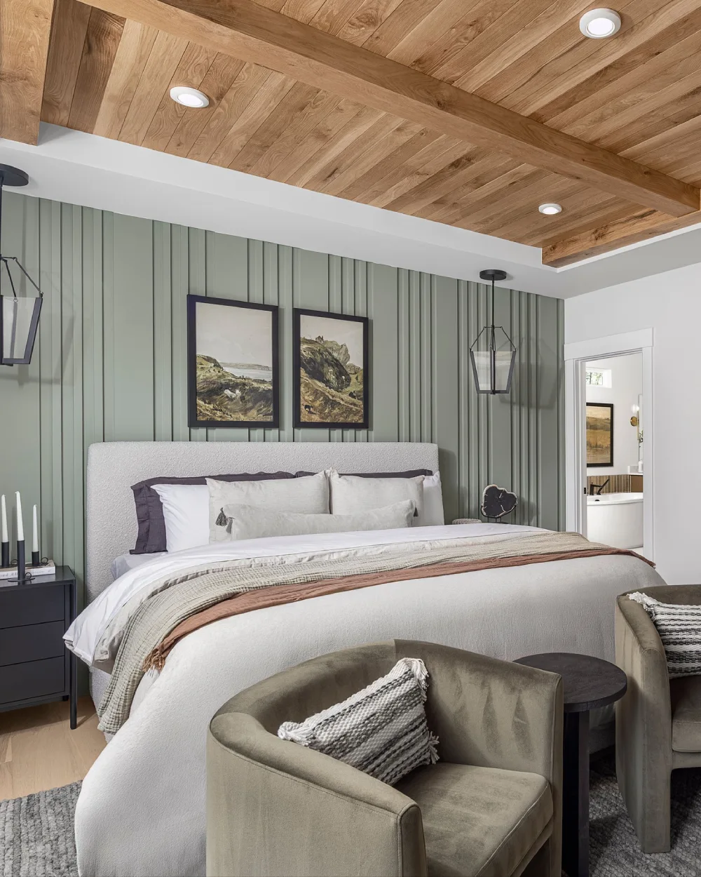 Muted green vertical paneled bedroom wall with a white upholstered bed, natural wood plank ceiling, framed landscape art, and olive green accent chairs.