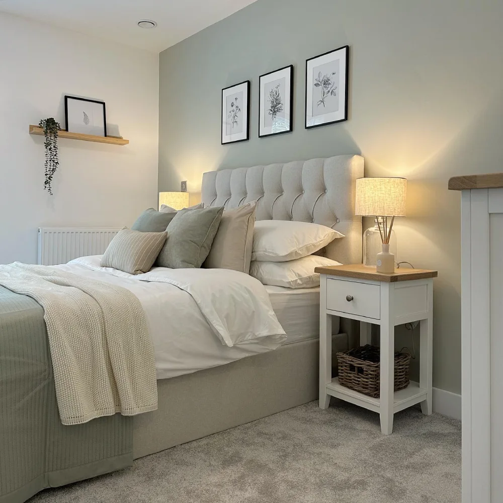 Soft green bedroom with a light gray upholstered headboard, white bedding, botanical artwork, and warm bedside lighting