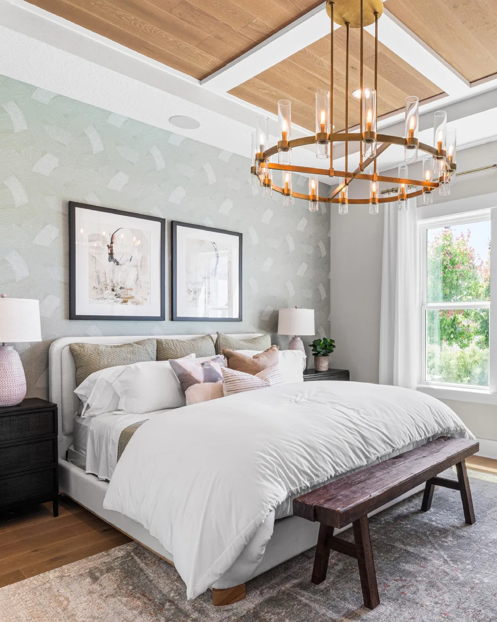 Light green patterned wallpaper behind a white upholstered bed with white bedding, blush accent pillows, a wooden ceiling, and a brass chandelier.