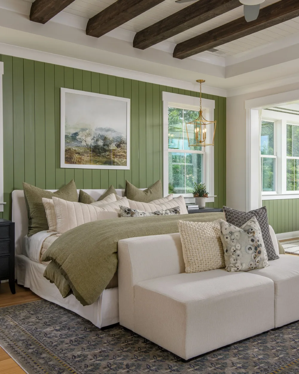 Olive green vertical paneled accent wall with white bedding, layered olive and cream pillows, dark exposed ceiling beams, and a cream upholstered bench.