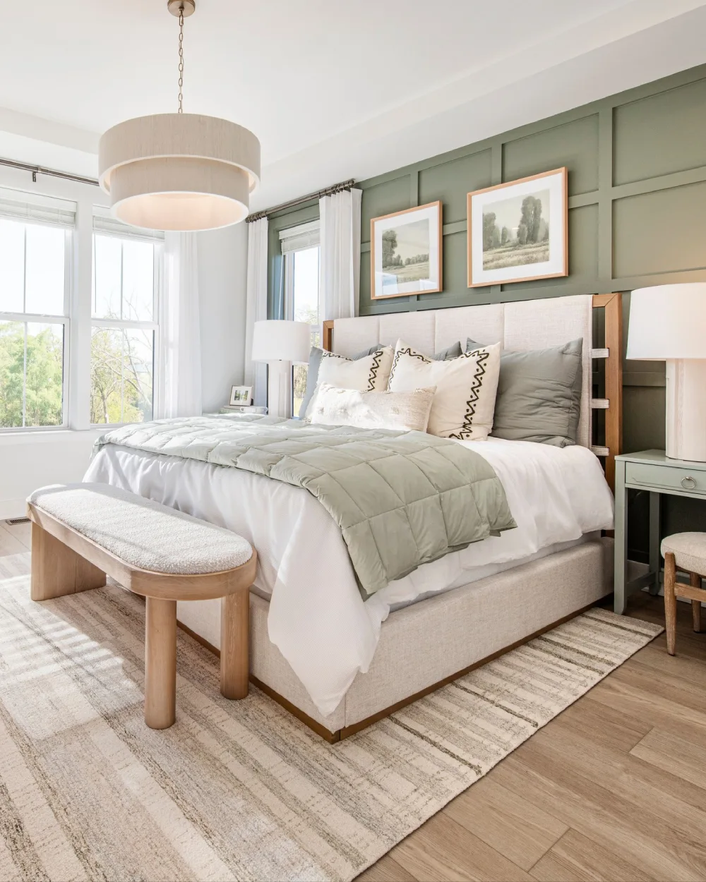 Muted green board and batten accent wall behind a beige upholstered bed, layered white and sage green bedding, light wood bench, and neutral striped rug.