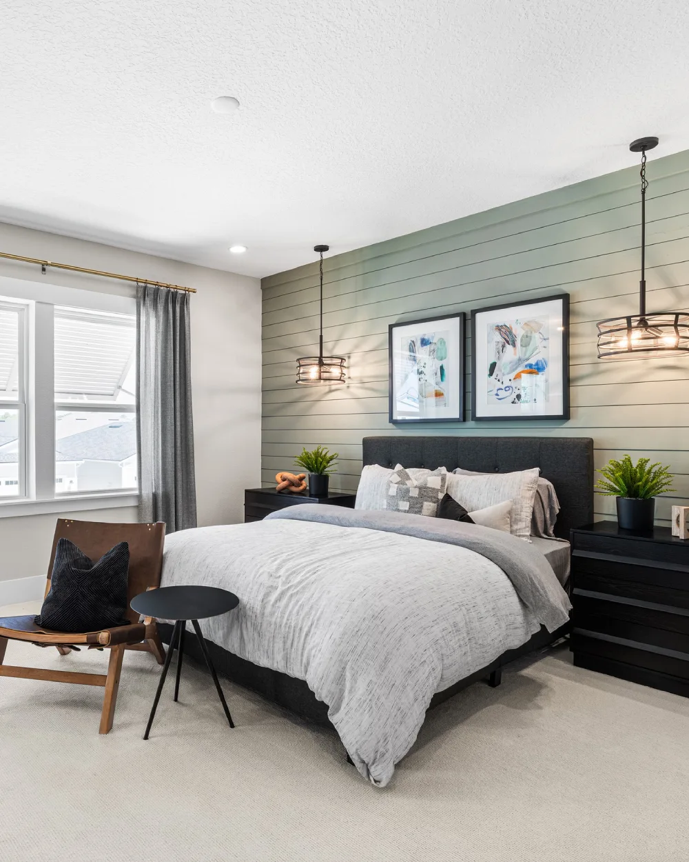 Sage green horizontal shiplap wall behind a dark upholstered bed with white bedding, black nightstands, framed artwork, and modern pendant lights.