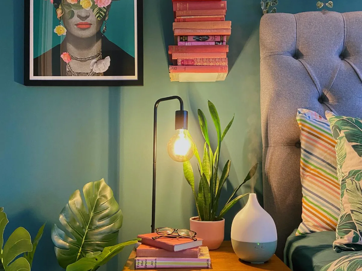 Green bedroom with teal walls, botanical artwork, leafy houseplants, a warm bedside lamp, and a gray upholstered bed with tropical leaf pillows.