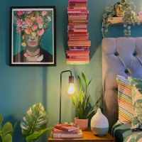 Green bedroom with teal walls, botanical artwork, leafy houseplants, a warm bedside lamp, and a gray upholstered bed with tropical leaf pillows.