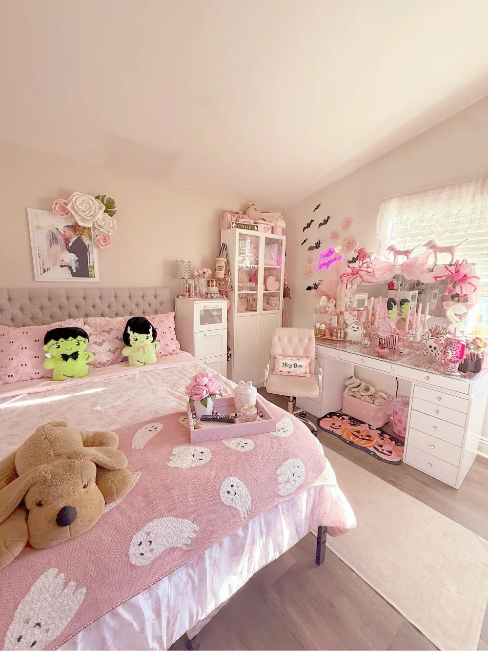 Pink and white bedroom with a tufted headboard, ghost-print pink blanket, plush toys, white vanity, and playful Halloween decor