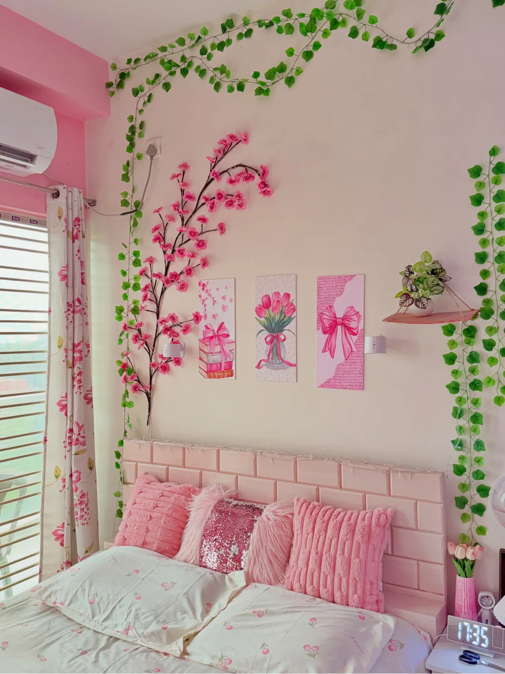 Pink bedroom with floral wall prints, trailing green vines, pink pillows, and a blush paneled headboard