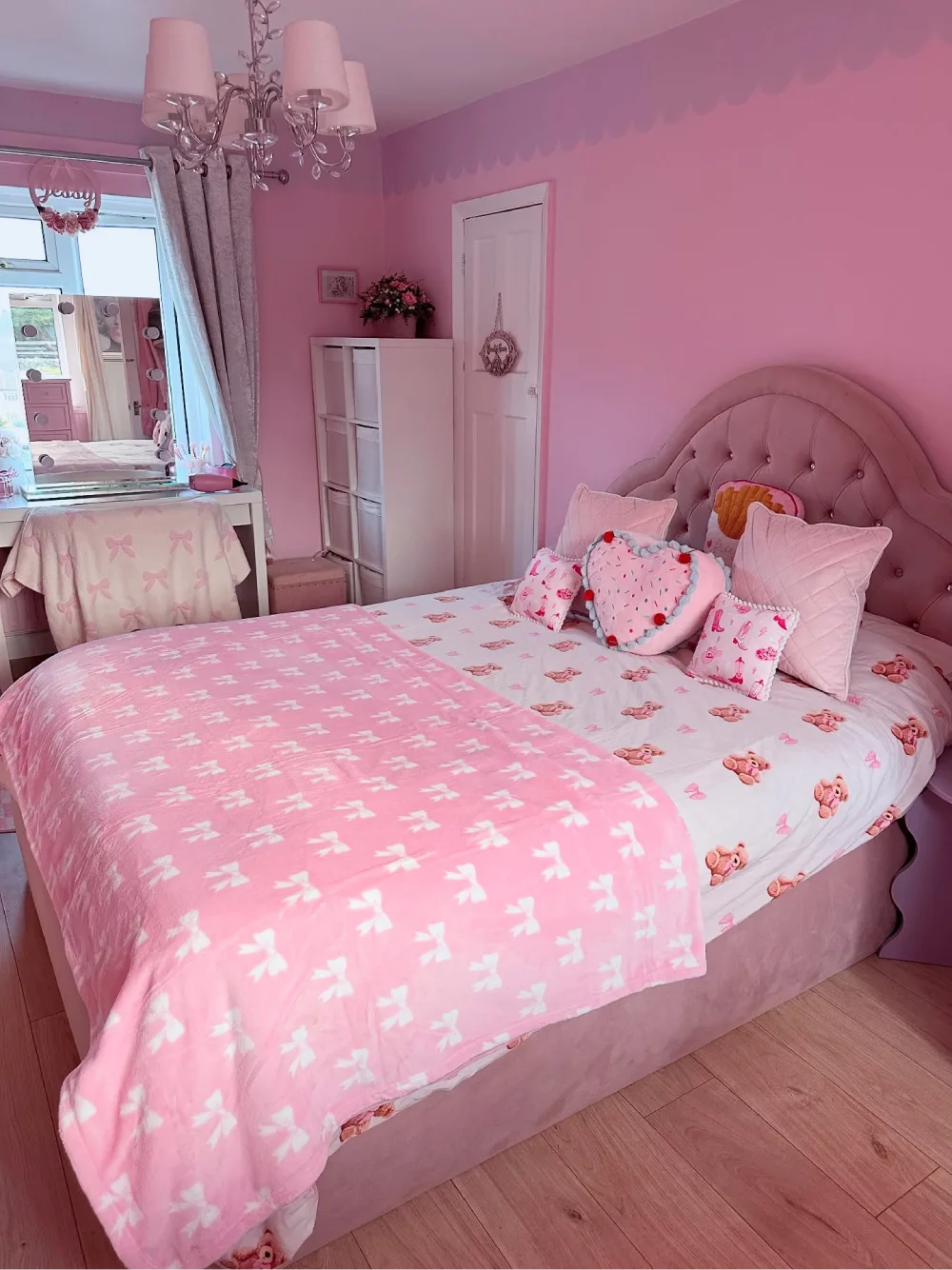 Bright pink bedroom with pink walls, a tufted upholstered headboard, floral bedding, bow-print blankets, and a white vanity