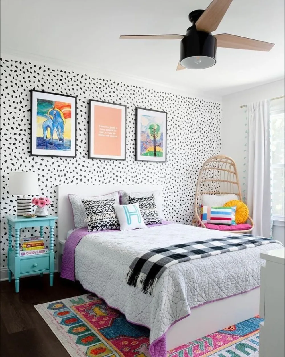 Bedroom with black polka dot wallpaper, colorful framed artwork, turquoise bedside table, purple bedding accents, and patterned rugs