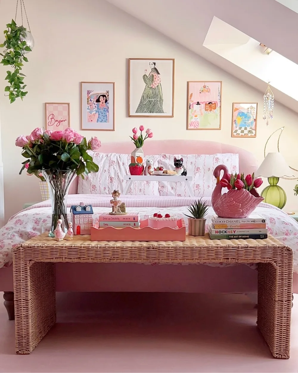 Romantic pink bedroom with a curved blush headboard, floral bedding, framed artwork, fresh flowers, and a woven wicker table