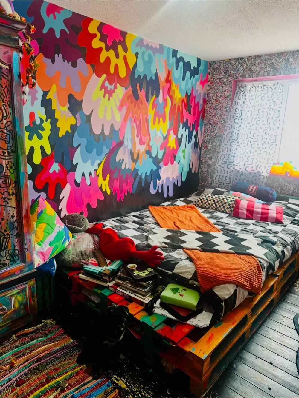 Eclectic bedroom featuring a vibrant abstract mural, black and white geometric bedding, colorful pillows, and stacked books