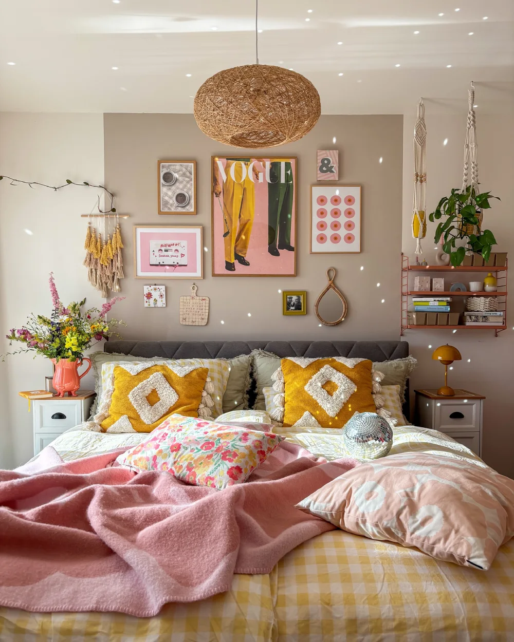 Cozy bedroom with a neutral upholstered headboard, mustard yellow pillows, a soft pink blanket, floral bedding, and colorful gallery wall