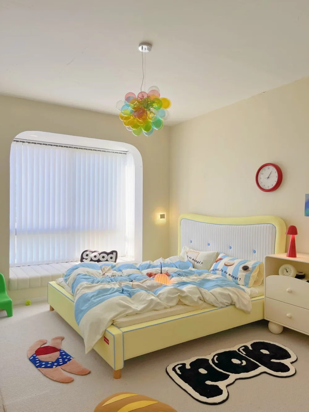 Pastel yellow dopamine decor bedroom with blue and white striped bedding, a colorful bubble chandelier, playful rugs, and a red bedside lamp