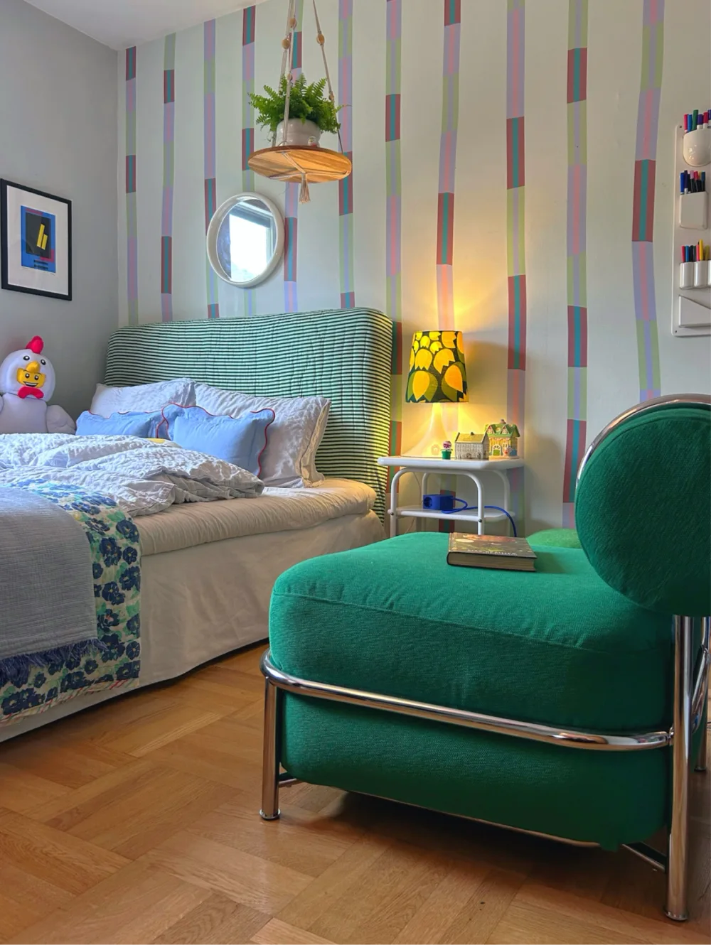 Bedroom with multicolored vertical striped wallpaper, a green upholstered chair, striped headboard, and cheerful yellow table lamp