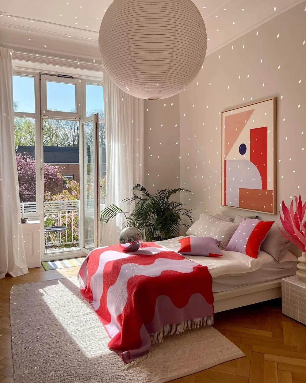 Bright bedroom with red and pink bedding, abstract wall art, an oversized paper pendant light, and sheer curtains framing tall windows.