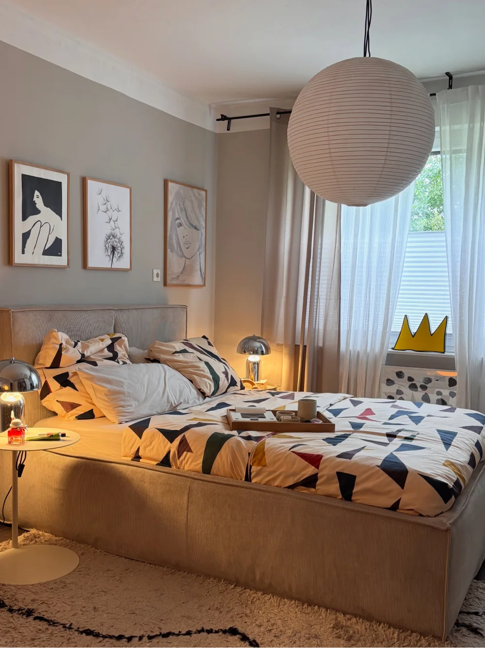 Bedroom with geometric patterned bedding, a beige upholstered bed, minimalist wall art, and a large paper pendant light