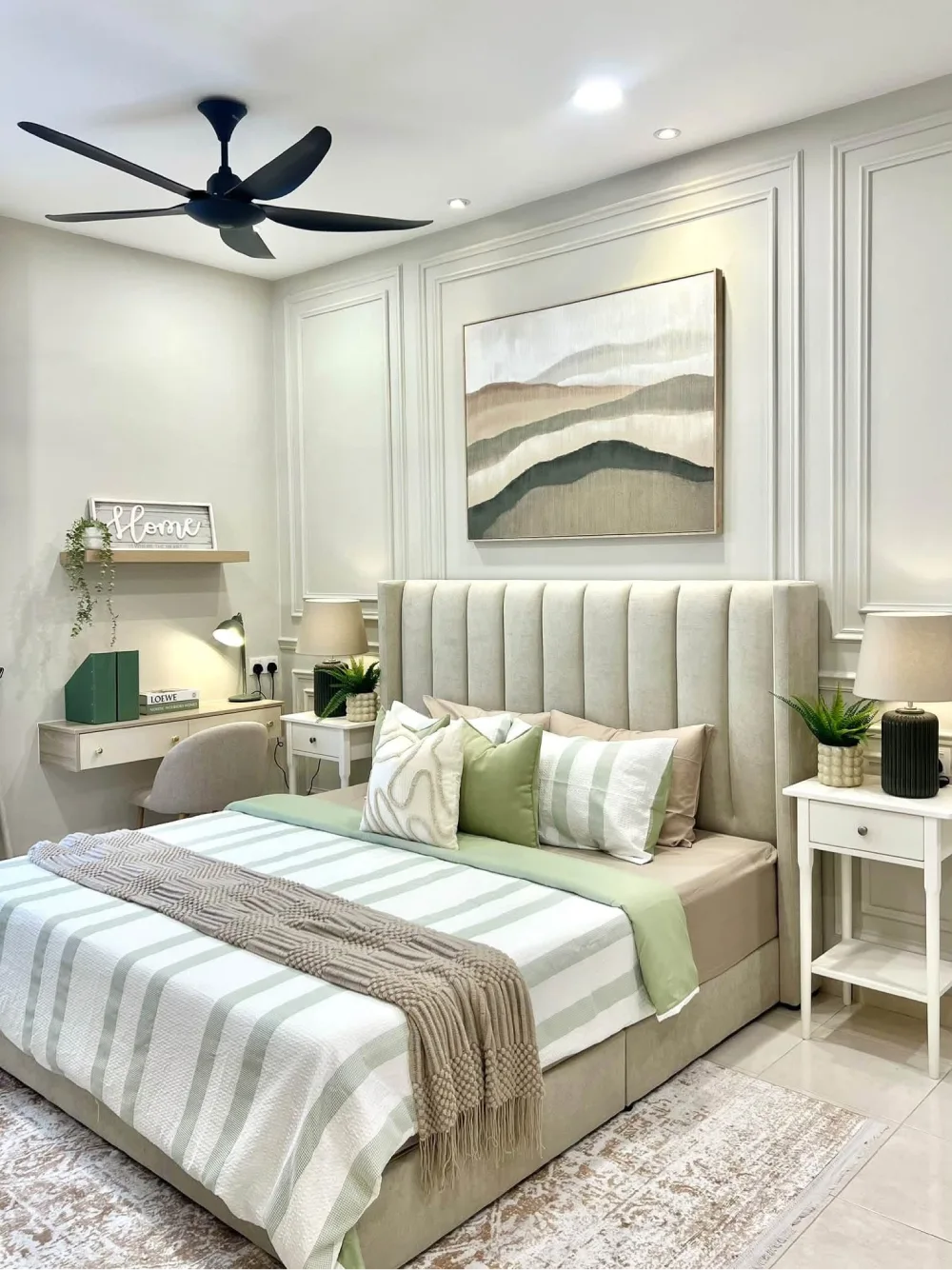 Elegant neutral bedroom with a tall beige upholstered headboard, white paneled walls, sage green bedding, matching nightstands, and landscape artwork.