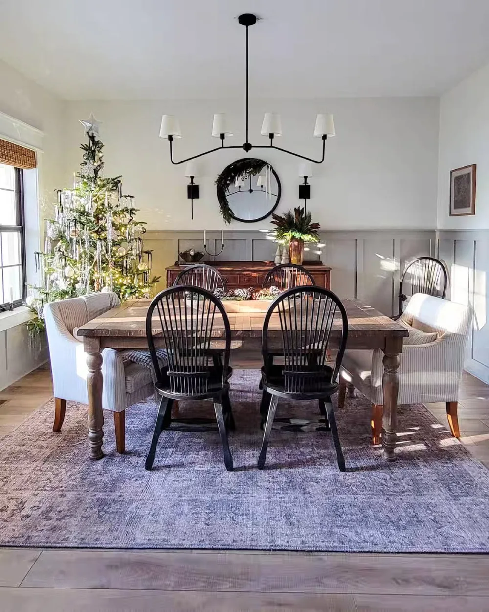Farmhouse winter dining room with a rustic wood table, black spindle chairs, greenery, and a Christmas tree