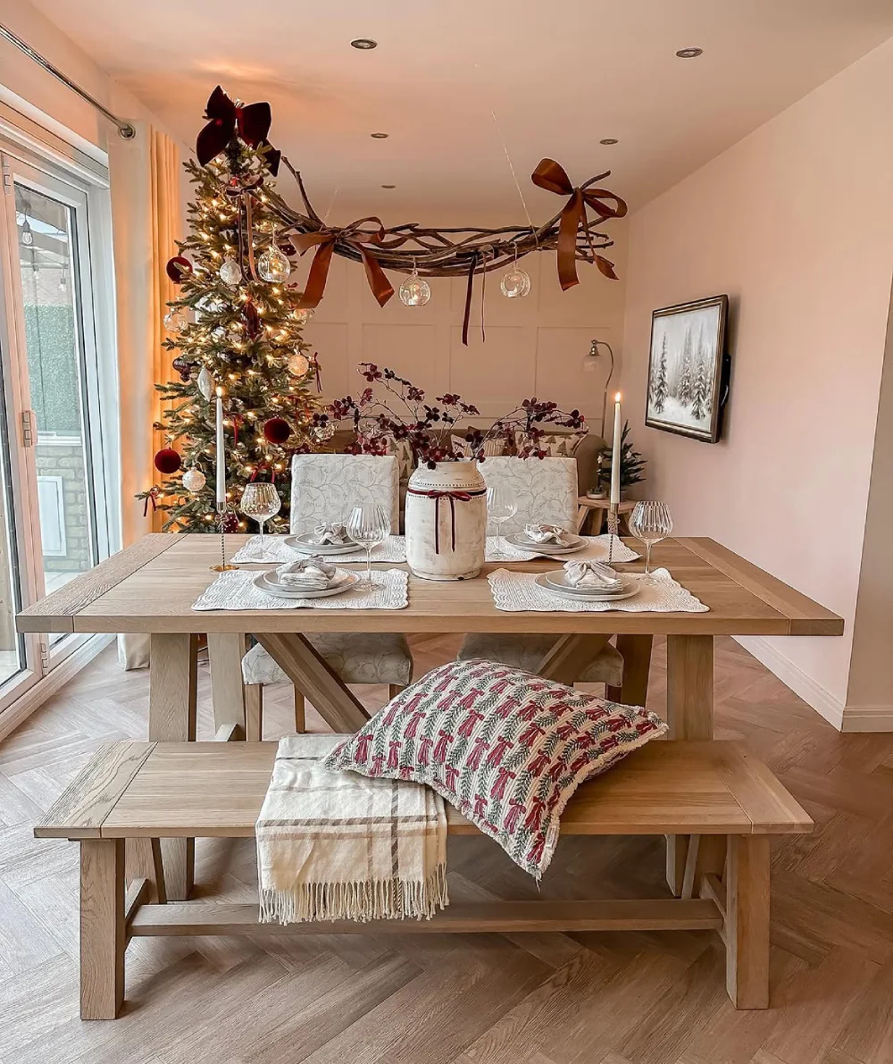 Warm winter dining room with a rustic wood table, Christmas tree, candles, and a cozy patterned pillow