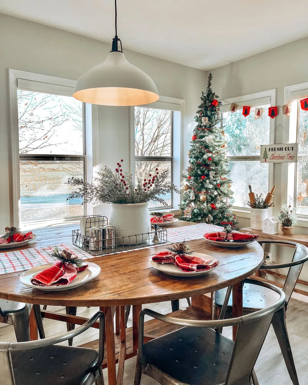 Bright winter dining nook with a round wooden table, red napkins, greenery, and a small Christmas tree