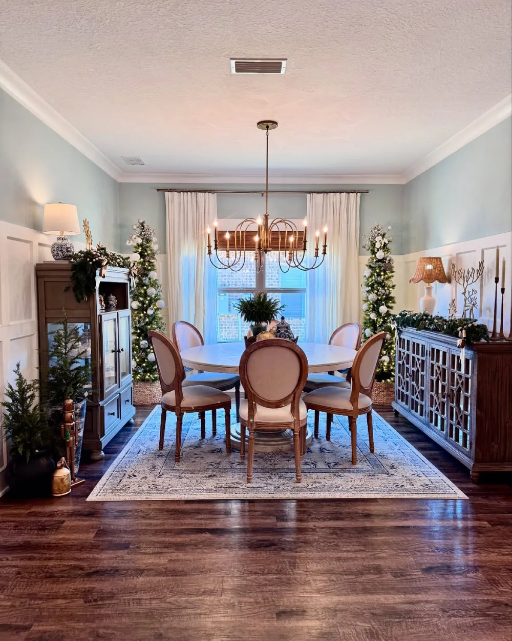 Classic winter dining room with a round table, traditional chairs, Christmas trees, and greenery