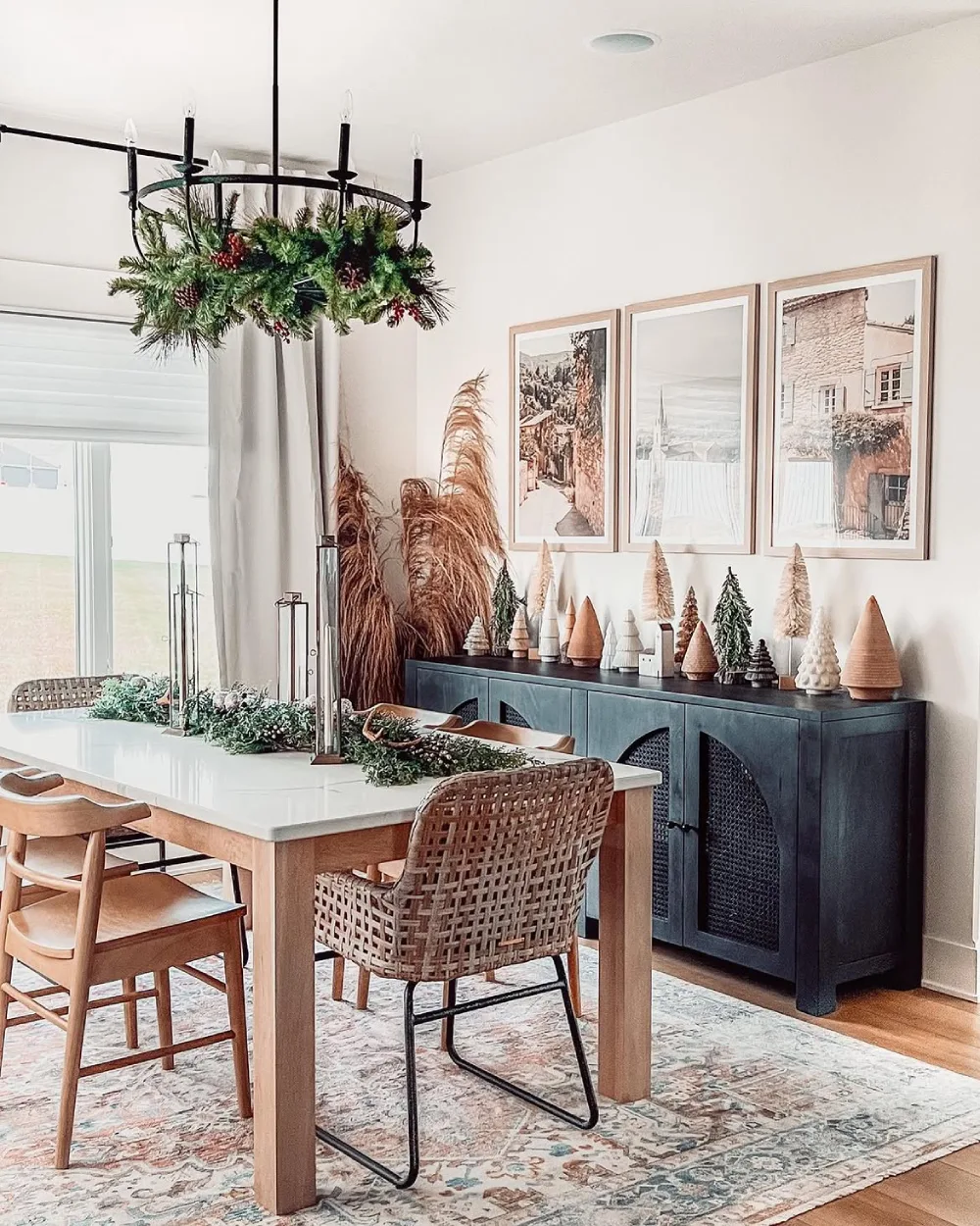 Bright winter dining room with a white table, woven chairs, evergreen garland, and mini decorative trees on a sideboard