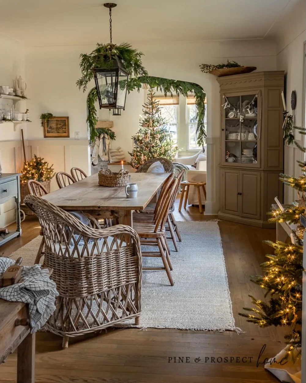 Warm farmhouse dining room with a rustic wood table, wicker chairs, greenery, and Christmas trees