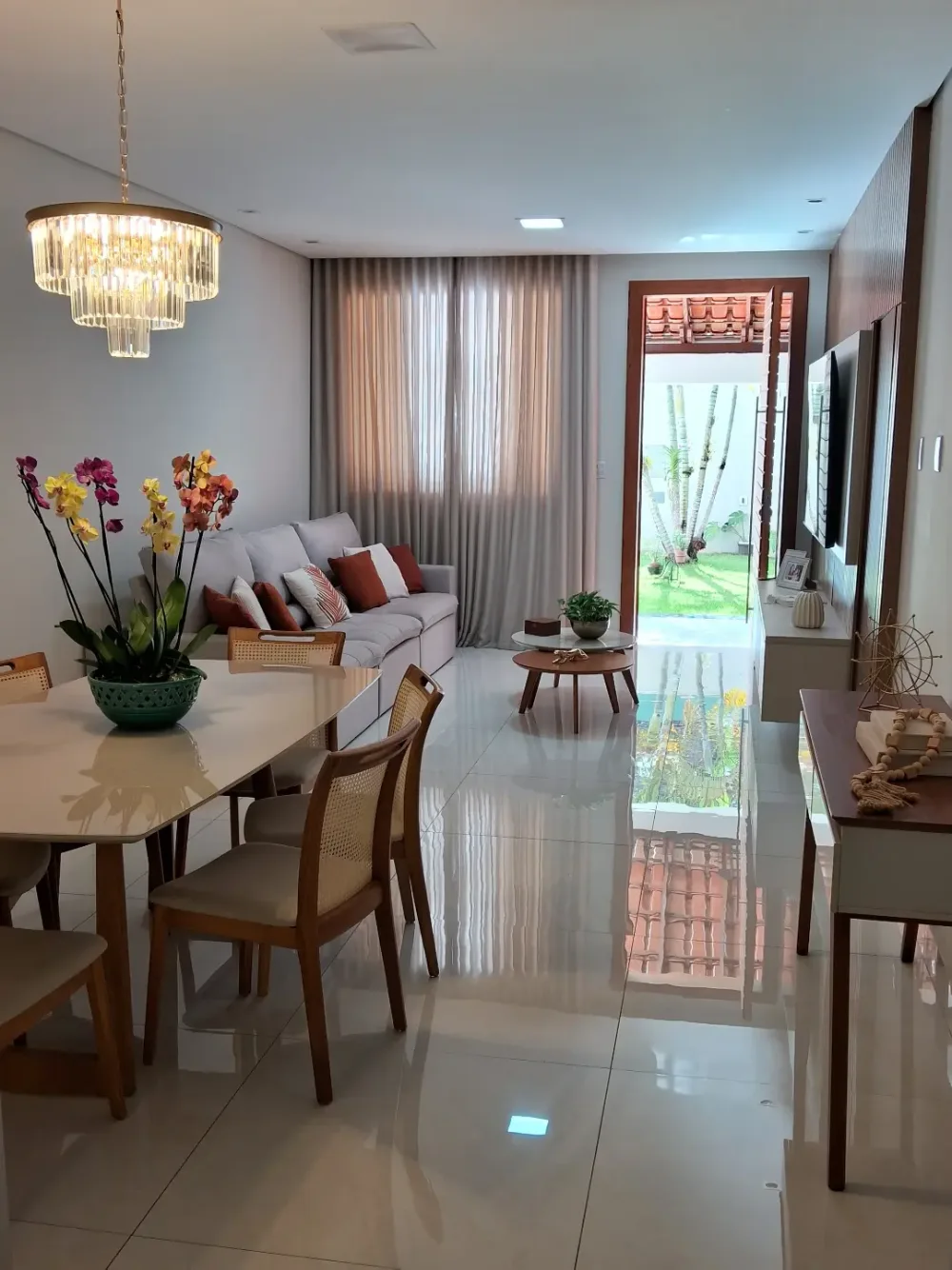 Open dining and living room with a white dining table, wooden chairs, crystal chandelier, and large windows