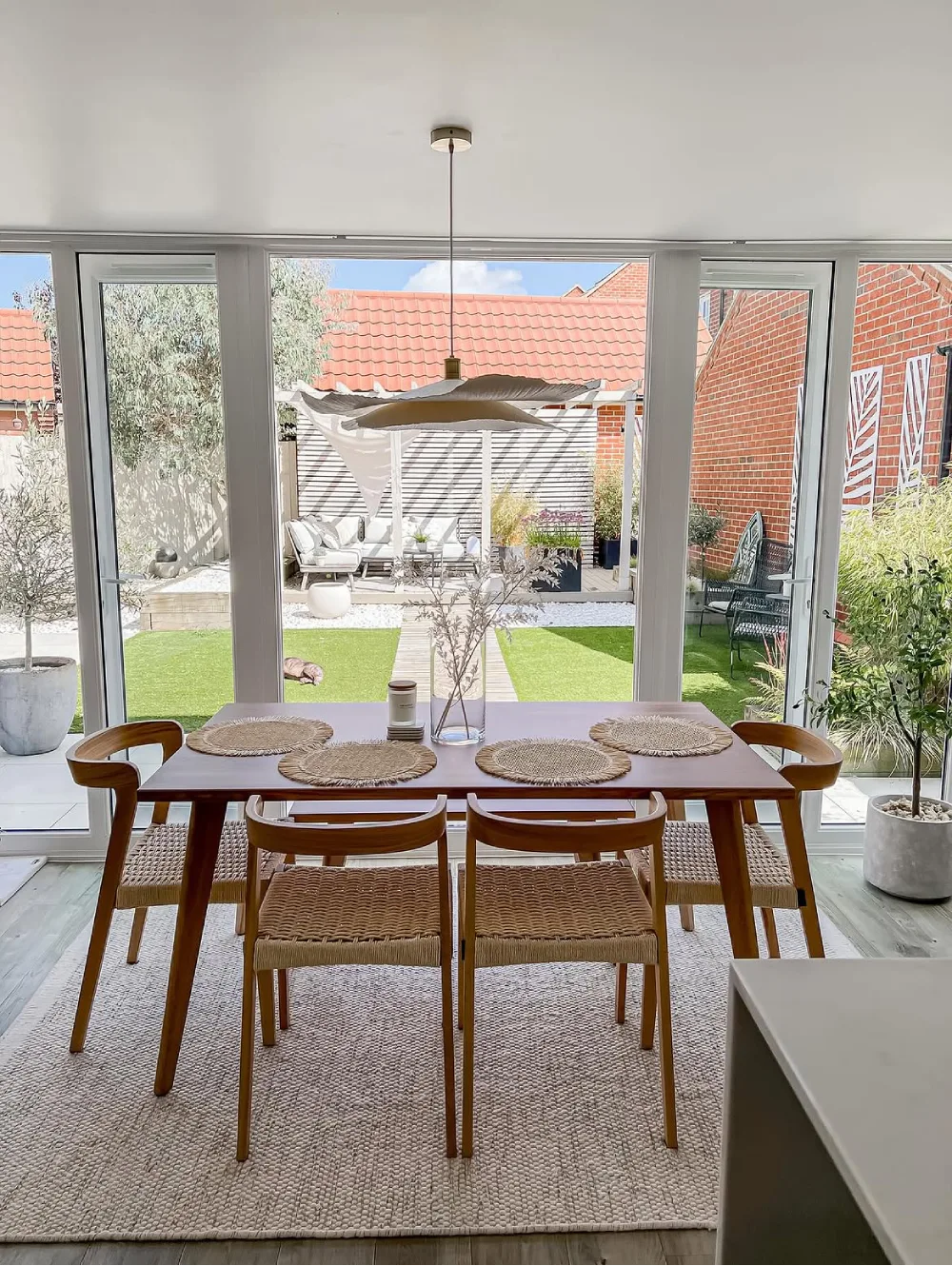 Bright dining room with a wood table, woven chairs, pendant light, and large glass doors overlooking a garden