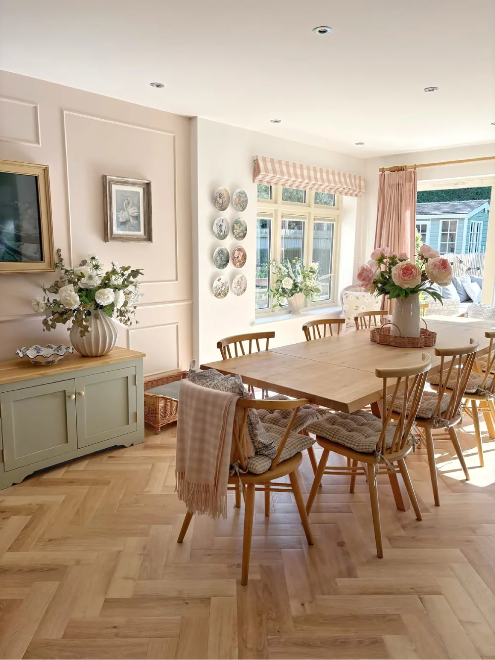 Cottage-style dining room with a long wood table, spindle-back chairs, floral arrangements, and soft pink curtains