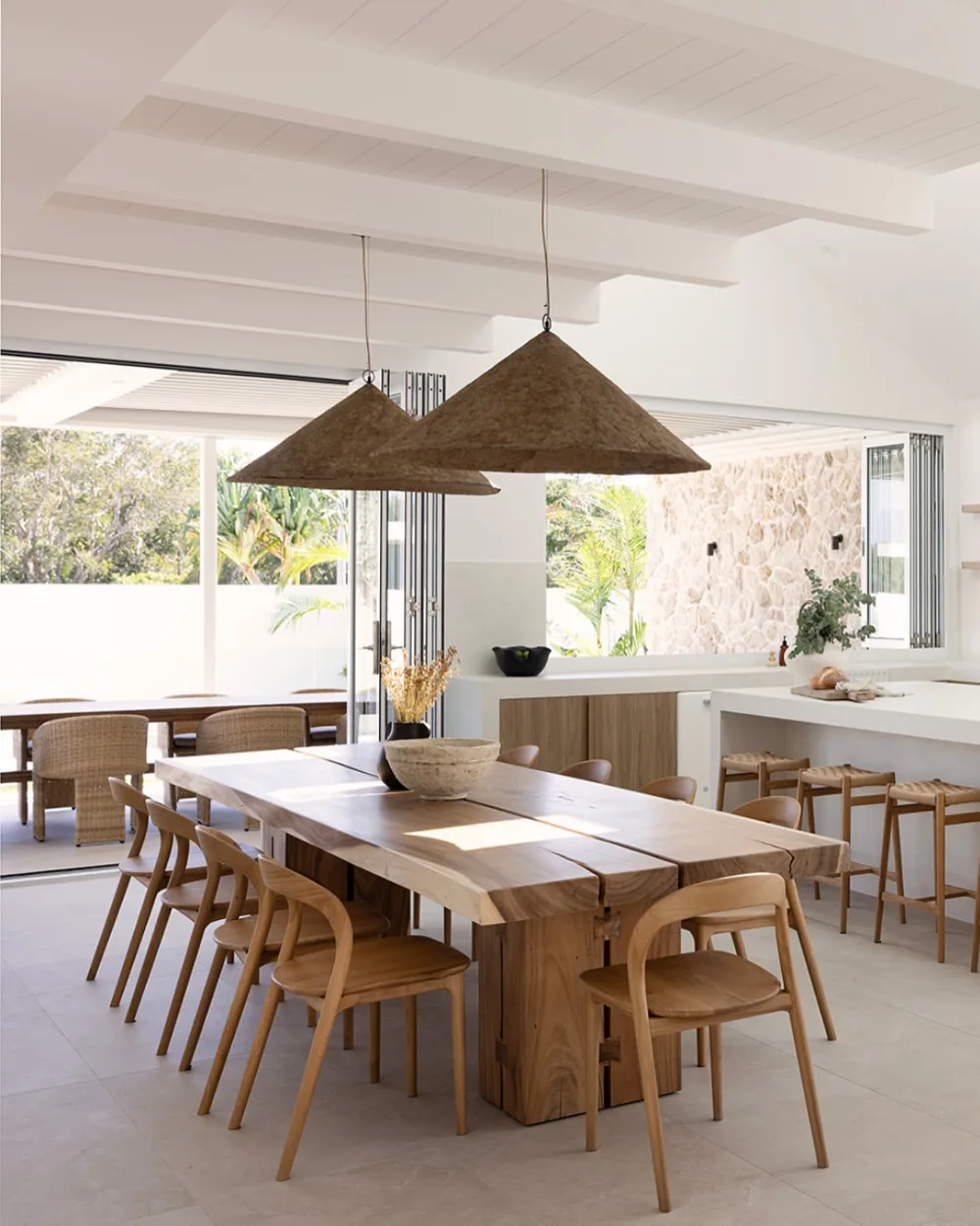Open dining area with a large wood table, woven pendant lights, wooden chairs, and a bright white kitchen